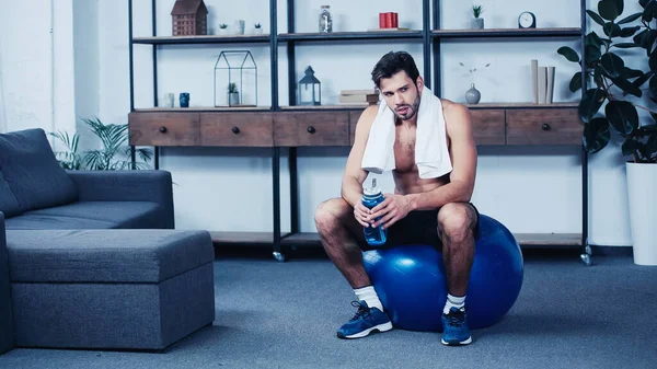 Deportista exhausto con toalla sentado en la pelota de fitness con botella de deporte - foto de stock