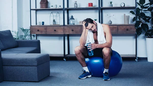 Уставший спортсмен с полотенцем сидит на фитнес-мяч со спортивной бутылкой — стоковое фото