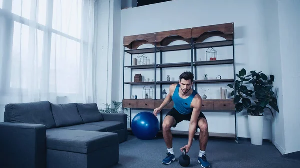 Barbudo joven deportista en camiseta azul ejercicio con kettlebell en casa - foto de stock
