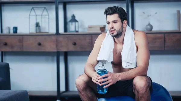 Tired young sportsman with towel sitting on fitness ball and holding sports bottle — Fotografia de Stock