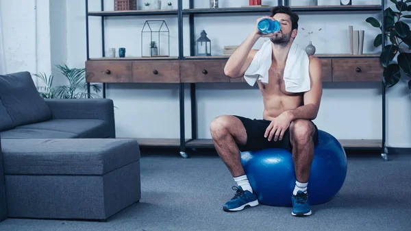 Full length of tired sportsman with towel sitting on fitness ball and holding sports bottle while drinking water — стоковое фото