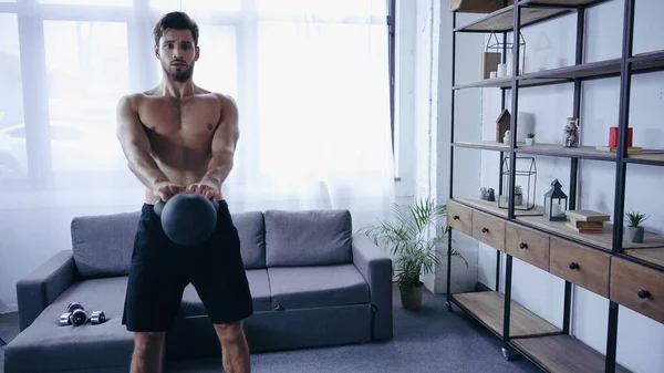 Young muscular sportsman in shorts training with heavy kettlebell near sofa — стоковое фото