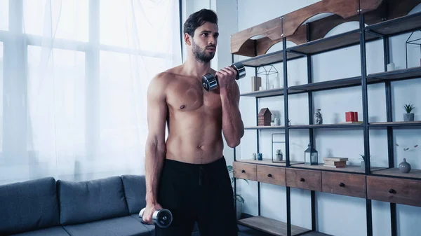 Homem muscular em treinamento de shorts com halteres pesados perto de rack — Fotografia de Stock