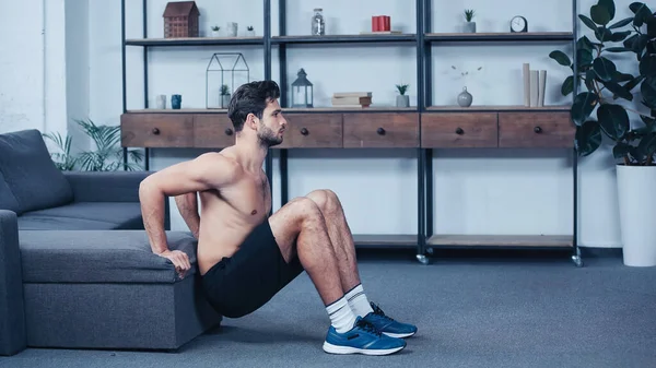 Vista laterale dello sportivo senza maglietta in pantaloncini accovacciato vicino al divano a casa — Foto stock