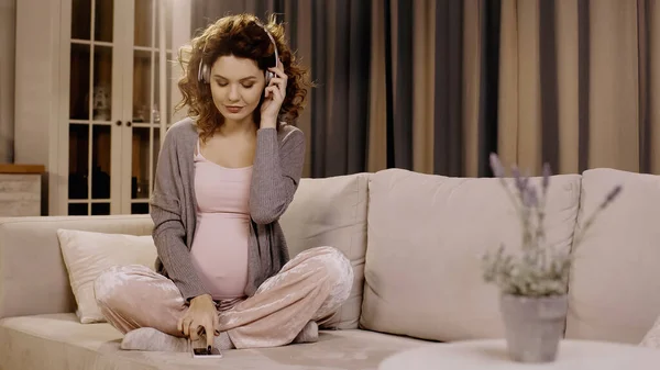 Pregnant woman in headphones using smartphone on couch at home — Stock Photo
