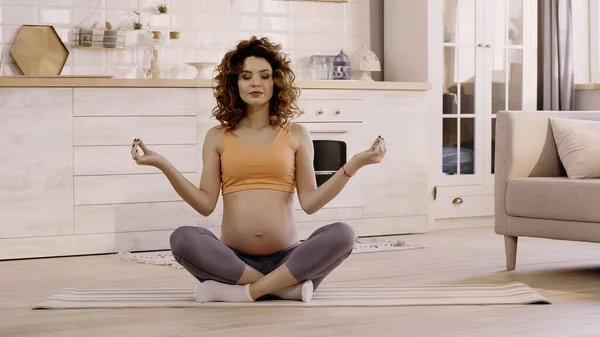 Mujer embarazada en ropa deportiva sentada en postura de yoga en la esterilla en casa - foto de stock