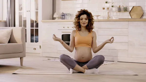 Mujer embarazada sonriente con los ojos cerrados sentada en postura de yoga en casa - foto de stock