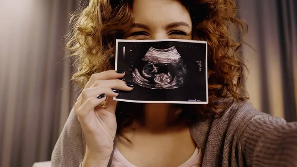 Lockige Frau bedeckt Gesicht mit Ultraschallbild des Babys zu Hause — Stockfoto