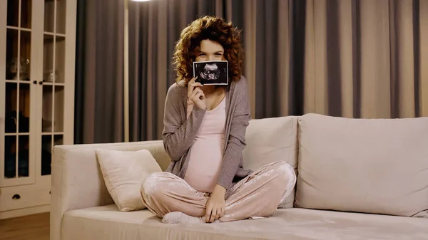 Pregnant woman covering face with ultrasound scan of baby at home — стоковое фото