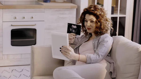 Pregnant woman showing ultrasound scan of baby during video call on digital tablet — стоковое фото