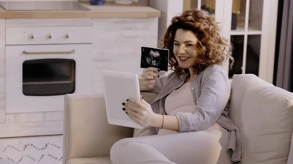 Happy pregnant woman with ultrasound scan of baby having video call on digital tablet at home — Foto stock