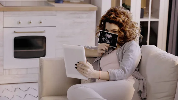 Pregnant woman holding ultrasound scan of baby during video call on digital tablet on couch — Fotografia de Stock