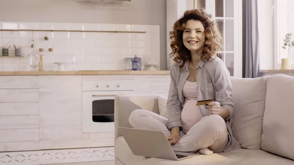 Mujer embarazada sosteniendo la tarjeta de crédito y sonriendo a la cámara cerca de la computadora portátil en el sofá - foto de stock