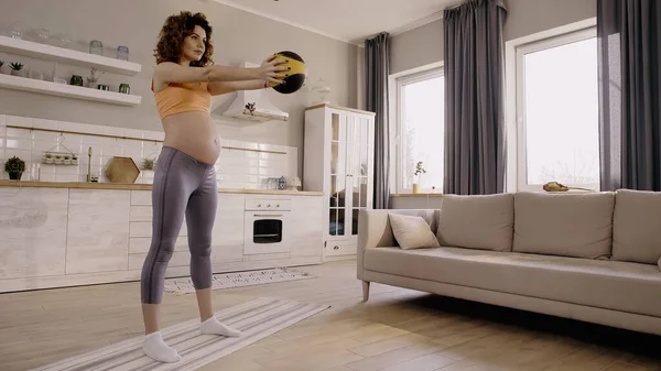 Curly pregnant sportswoman exercising with slam ball on fitness mat at home — Stock Photo
