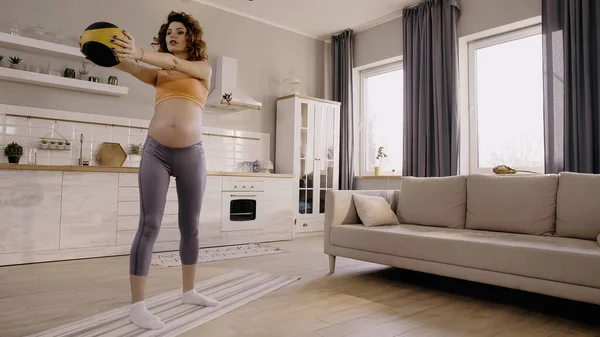 Sportive enceinte tenant le ballon de slam pendant l'entraînement sur tapis de fitness à la maison — Photo de stock