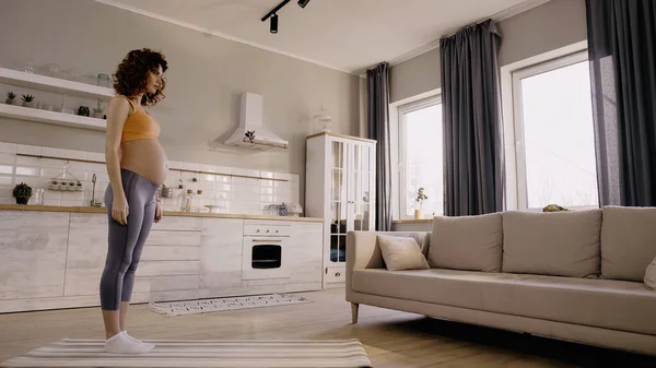 Vue latérale d'une jeune sportive enceinte debout sur un tapis de fitness à la maison — Photo de stock