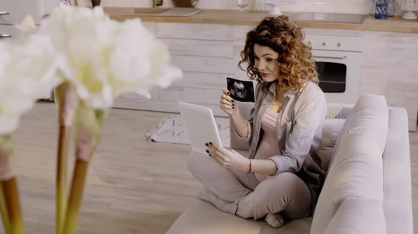 Femme enceinte montrant échographie du bébé lors d'un appel vidéo sur tablette numérique sur le canapé dans le salon — Photo de stock