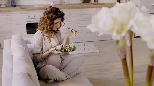 Curly pregnant woman holding salad and fork on couch at home — стоковое фото