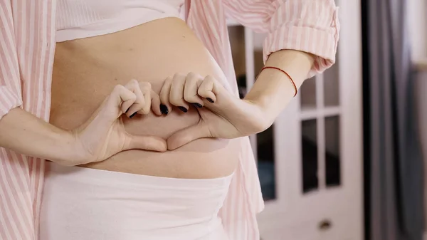 Ausgeschnittene Ansicht einer schwangeren Frau mit Herzzeichen in der Nähe des Bauches zu Hause — Stockfoto