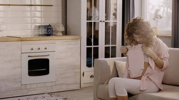 Curly pregnant woman holding baby clothes on couch in kitchen — Fotografia de Stock