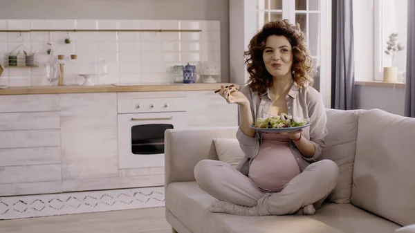 Happy pregnant woman holding plate with fresh salad and fork on couch — Fotografia de Stock