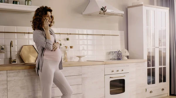 Curly pregnant woman talking on smartphone near worktop in kitchen — Stock Photo