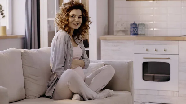 Mujer embarazada sonriente mirando a la cámara mientras está sentada en el sofá en casa - foto de stock