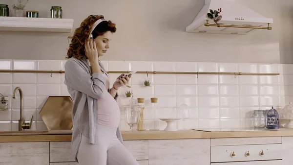 Mujer embarazada joven escuchando música en auriculares y sosteniendo el teléfono inteligente en la cocina - foto de stock