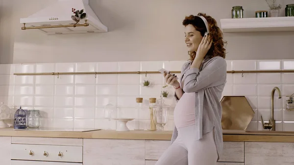 Cheerful pregnant woman using headphones and cellphone in kitchen — стоковое фото