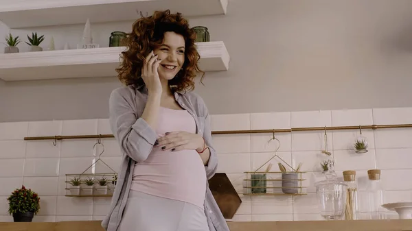 Sonriendo mujer embarazada hablando en el teléfono inteligente en la cocina en casa - foto de stock