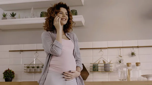 Mujer embarazada sonriente tocando el vientre mientras habla en el teléfono inteligente en casa - foto de stock