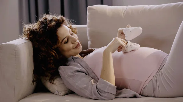 Sourire femme enceinte tenant des chaussons de bébé tout en étant couché sur le canapé — Photo de stock