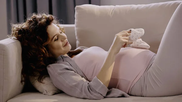 Curly pregnant woman holding baby booties near belly on couch — стоковое фото