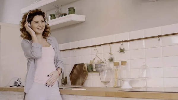 Happy pregnant woman listening music in headphones near cellphone on worktop in kitchen — стоковое фото