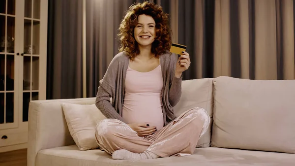 Mujer embarazada positiva sosteniendo la tarjeta de crédito y sonriendo a la cámara en casa - foto de stock