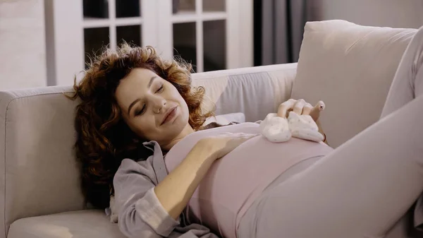 Pleased pregnant woman holding baby booties near belly on couch — Stock Photo