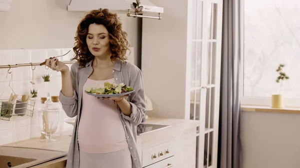 Радостная беременная женщина держит свежий салат и вилку на кухне — стоковое фото