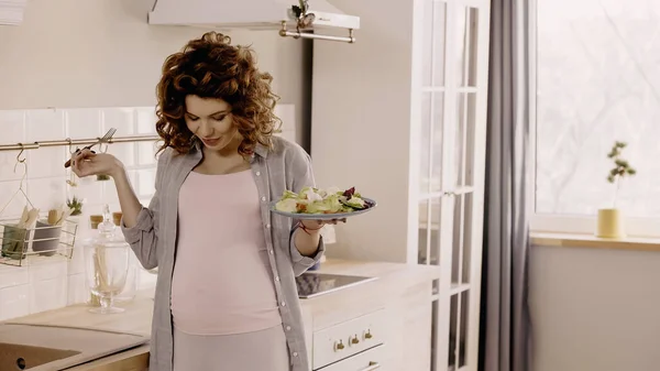 Curly pregnant woman holding fork and salad while looking at belly in kitchen — Stockfoto