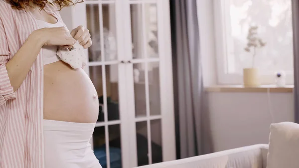 Vista recortada de la mujer embarazada sosteniendo botines de bebé en la sala de estar - foto de stock