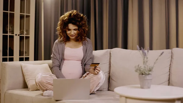 Femme enceinte tenant une carte de crédit et utilisant un ordinateur portable sur le canapé — Photo de stock