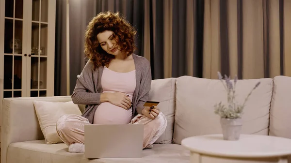 Mujer embarazada joven tocando el vientre y la celebración de la tarjeta de crédito cerca de la computadora portátil en el sofá en casa - foto de stock