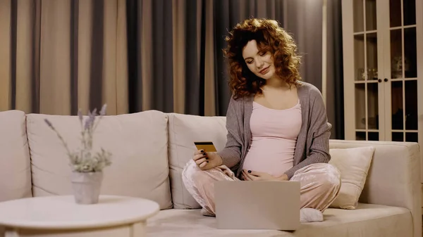 Беременная женщина с кредиткой рядом с ноутбуком и размытым растением на диване дома — стоковое фото