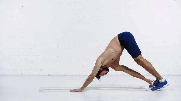 Vue latérale d'un sportif torse nu s'étirant vers l'arrière sur un tapis de fitness près d'un mur de briques blanches — Photo de stock