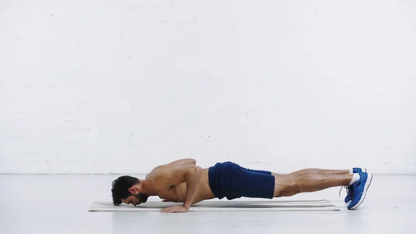 Sin camisa deportista haciendo tablón en la estera de fitness cerca de la pared de ladrillo blanco - foto de stock