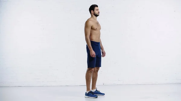 Full length of shirtless sportsman in shorts standing near white brick wall — Stockfoto