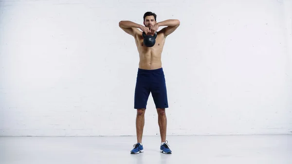 Full length of muscular sportsman exercising with heavy kettlebell near white brick wall — стоковое фото