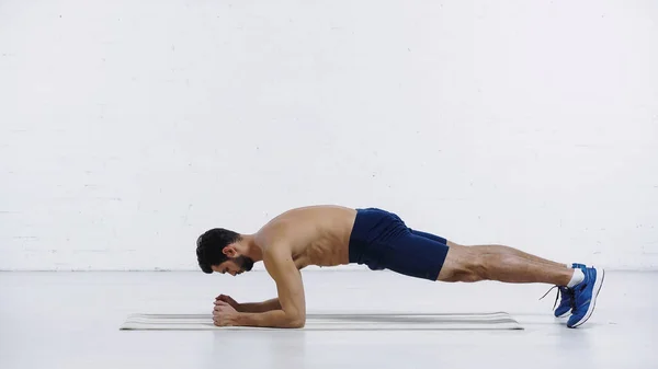 Vista lateral del deportista sin camisa haciendo ejercicio de tabla en la estera de fitness cerca de la pared de ladrillo blanco - foto de stock