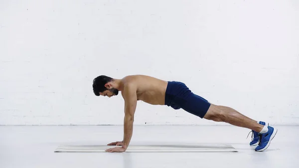 Vista lateral del deportista sin camisa en pantalones cortos haciendo tablón en la estera de fitness cerca de la pared de ladrillo blanco - foto de stock