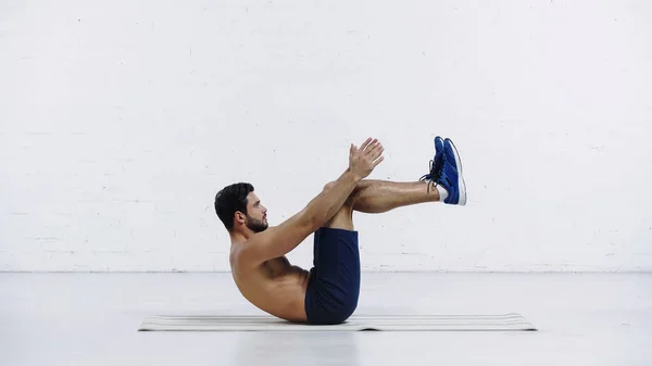 Vista lateral del deportista barbudo en pantalones cortos haciendo ejercicio en la estera de fitness cerca de la pared de ladrillo blanco - foto de stock