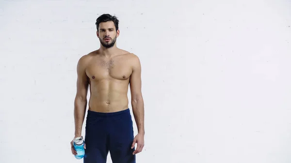 Bearded sportsman in shorts holding blue sports bottle near white brick wall — Stock Photo